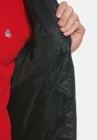 Schwarze Jackeninnenseite mit glatter Textur und Zipper-Tasche, getragen über einem roten Strickoberteil mit einem kleinen Pinguin-Logo.