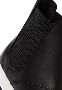 Zapato negro de piel sin cordones con panel lateral elástico texturizado y suela blanca, mostrando detalles traseros y laterales.