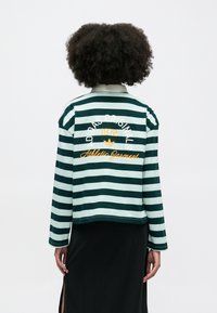 Zaļi un gaiši zili svītrota garām piedurknēm krekls ar apkakles izgriezumu, kurā ir balti un oranži uzraksti "Adidas Original 1972 Athletic Garment."