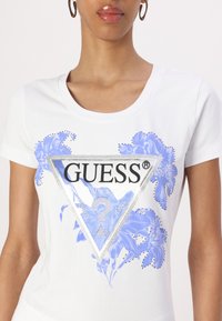 Bílý tričko s modrým květinovým vzorem uvnitř trojúhelníkového loga, s detaily z kamínků a černým textem "GUESS" a "U.S.A."