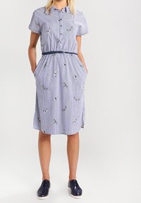 Kvinna som bär en blå och vit randig knälång klänning med fjärilsmönster, korta ärmar, marinblått bälte och marinblå snörskor.