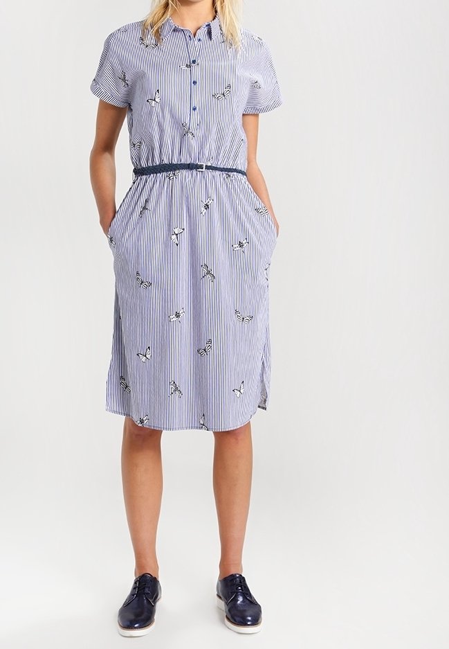 Kvinna som bär en blå och vit randig knälång klänning med fjärilsmönster, korta ärmar, marinblått bälte och marinblå snörskor.