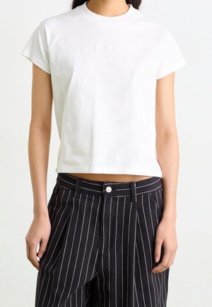 Femme portant un t-shirt court blanc et un pantalon noir taille haute à fines rayures blanches, debout devant un fond uni.