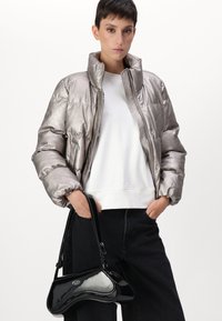 Femme portant une doudoune argent métallique, top blanc, pantalon noir, tenant un petit sac à main en cuir verni noir avec une sangle courte.