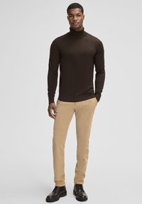 Maglione dolcevita marrone abbinato a pantaloni in velluto a coste beige. Il modello indossa stivali neri con lacci. Design semplice e aderente, texture liscia.