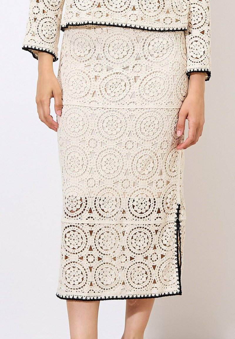 Jupe midi en dentelle crème avec des motifs circulaires, rehaussée de poignées noires. Présente une silhouette ajustée et un détail fendu sur le côté.