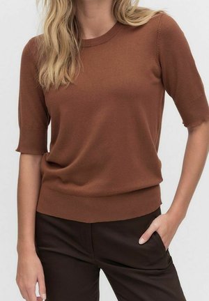 Femme portant un haut en maille à manches courtes marron et un pantalon marron foncé, avec une main dans la poche, sur un fond clair uni.