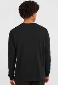 Young person with curly hair wearing a plain black long-sleeve shirt facing away against a white background.