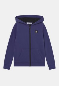 Sudadera morada con cremallera, forro negro, dos bolsillos frontales y un pequeño parche con logo. Material de algodón, corte estándar, capucha ajustable.