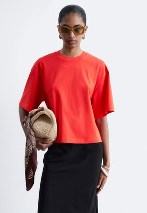 Femme portant un t-shirt oversize orange vif et une jupe noire, tenant un sac tissé avec une écharpe à motifs, portant des lunettes de soleil marron et des boucles d'oreilles créoles.