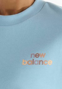 Close-up of blue sweatshirt with ribbed neckline and "new balance" logo in gradient orange and purple lettering on chest.