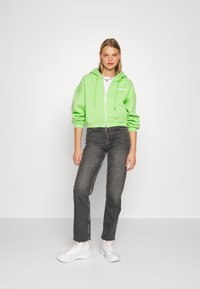 Moletom curto verde claro com fecho, apresentando um capuz com cordão e logotipo na frente. Combinado com calças jeans cinza de cintura alta e ténis brancos.