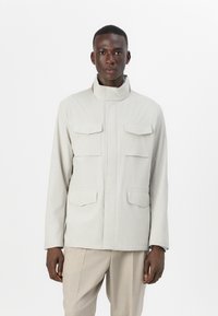 Light beige jacket with a high collar, featuring two front chest pockets and a flat design. Fabric appears smooth and lightweight.