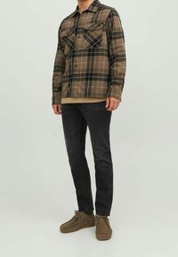 Chaqueta de cuadro marrón con dos bolsillos en el pecho, llevada sobre una camisa beige, combinada con pantalones vaqueros negros y zapatos marrones. Material de forro polar.