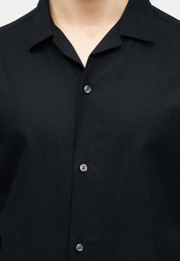 Κοντινό πλάνο ενός ατόμου που φοράει μαύρο πουκάμισο με κουμπιά και γιακά, με ορατά κουμπιά στη μπροστινή πλευρά.