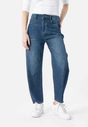 Person wearing blue straight-leg jeans and white sneakers, with one hand in pocket and white long-sleeve top.