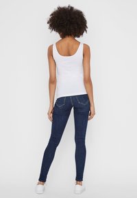 Débardeur blanc à bretelles larges et dos plongeant, associé à un jean skinny bleu foncé avec poches arrière et texture lisse.
