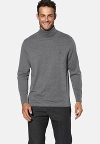 Grauer Rollkragenpullover mit langen Ärmeln, aus weichem Stoff. Mit einem dezenten Logo auf der Brust, kombiniert mit dunkler Jeans.