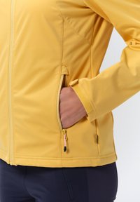 Żółta kurtka softshell z zamykanymi kieszeniami, elastycznymi mankietami i gładką teksturą. Posiada stojący kołnierz oraz przeszywane szwy.