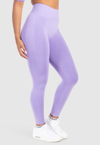 Leggings violettes taille haute avec une texture lisse et extensible, associés à des baskets blanches avec des accents noirs au niveau de la cheville.