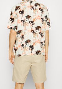 Camicia a maniche corte in tessuto leggero con uno sfondo acquerellato pastello e motivi di palme nere e arancioni. Abbinata a pantaloni corti beige.