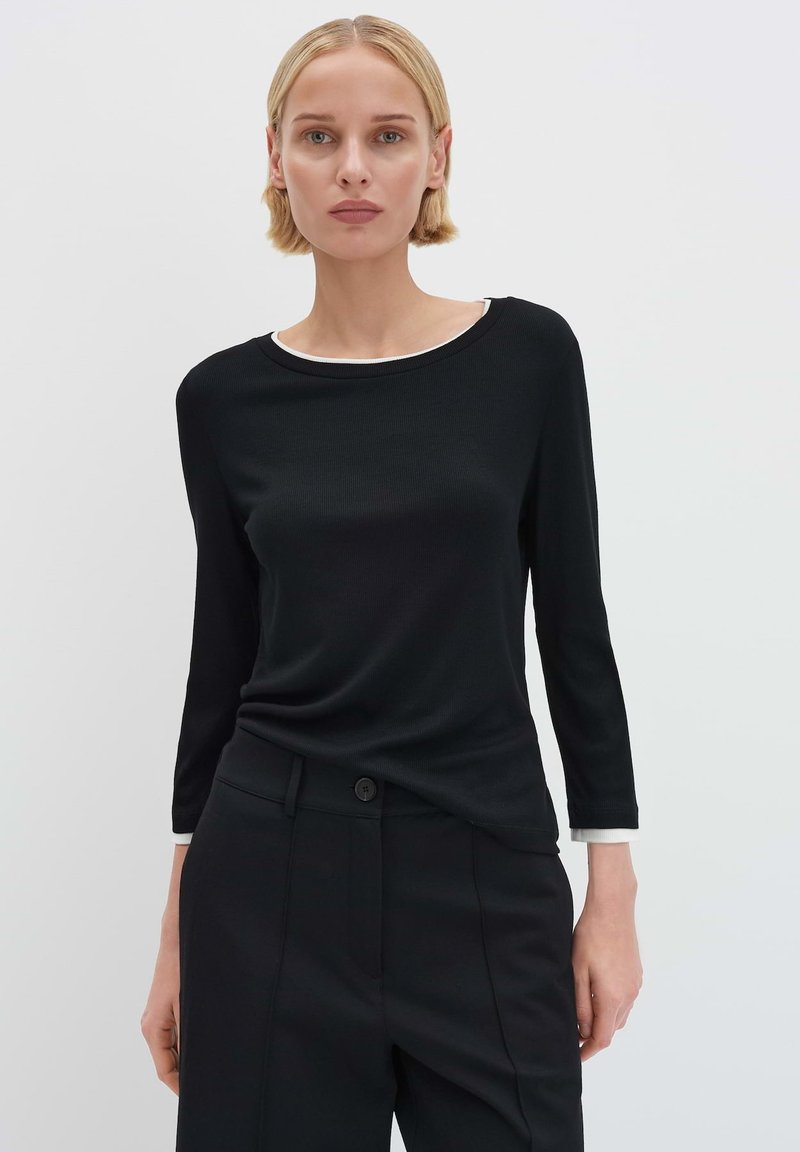 Schwarzes, geripptes Langarmshirt mit rundem Ausschnitt und weißem Besatz, kombiniert mit schwarzen, maßgeschneiderten Hosen. Der Stoff ist glatt und figurbetont.