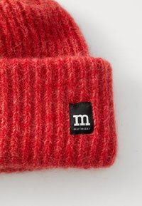 Red knit beanie made of textured wool, featuring a ribbed design and a black label with white lettering on the folded cuff.