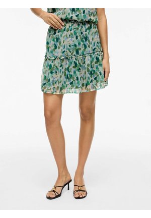 Femme portant une jupe courte verte et bleue à motifs avec des sandales à talons hauts noires et à lanières, debout sur un fond blanc.