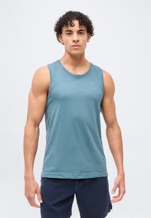 Młody mężczyzna o kręconych włosach, ubrany w niebieską koszulkę bez rękawów Nike i ciemne szorty, stojący przodem na jednolitym tle.