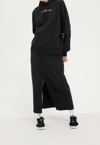 Czarna oversize'owa bluza z kapturem z różowym napisem, zestawiona z czarną długą spódnicą jeansową z przednim rozcięciem, noszona z czarnymi trampkami i białymi skarpetkami.