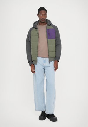 Green and gray padded jacket with a purple chest pocket, worn over a beige shirt and light blue jeans, complemented by black shoes.