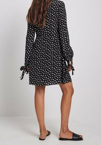 Schwarzes Kleid mit weißen Punkten und langen Ärmeln, das an den Manschetten mit Schleifen verziert ist. Leichter Stoff, etwa bis zur Mitte des Oberschenkels, kombiniert mit schwarzen Riemchensandalen.