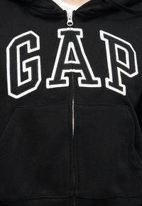 Sweat à capuche noir zippé avec de grandes lettres "GAP" blanches en contour sur la poitrine et des poches avant.