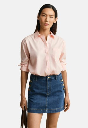 Femme aux longs cheveux bruns portant une chemise rose clair à boutons avec les manches retroussées et une jupe en jean bleue avec des boutons dorés sur les côtés.