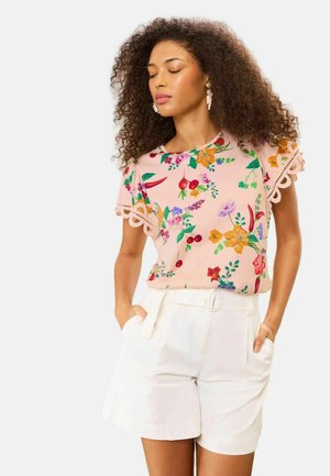 Naiste lokkis juustega, kes kannab roosat lillesusega pluusi pitsist varrukatega ning kõrge vöökohaga valgeid lühikesi pükse, käed taskutes.