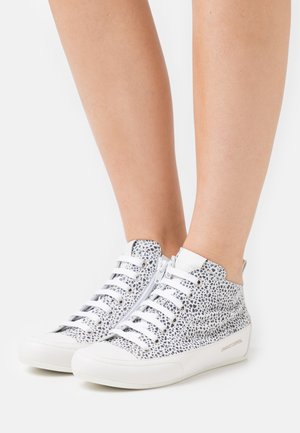 Baskets montantes blanches avec un motif tacheté noir, lacets blancs, fermeture éclair latérale, portées sur des jambes nues devant un fond uni.