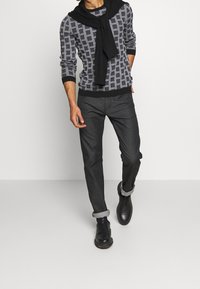 Mönstrad svart och vit tröja, mörkgrå byxor, svarta skor med uppvikt fåll, och en svart tröja draperad över axlarna.