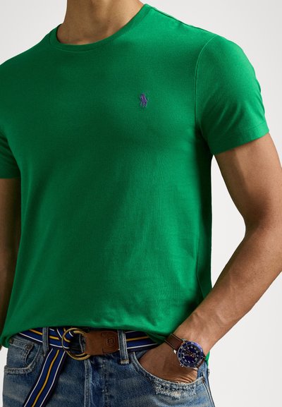 T-shirt de algodão verde com decote redondo e mangas curtas, apresentando um pequeno logotipo roxo. Cinto castanho com fivela decorativa e detalhes às riscas.