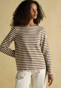 Beige Langarmshirt mit braunen horizontalen Streifen, weitem Ausschnitt und weichem Baumwollstoff. Getragen mit hellen Hosen.