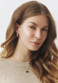Goldene Creolen und eine goldene Kettenhalskette mit einem rechteckigen Anhänger. Das Model hat lange, gewellte braune Haare und trägt einen hellbeigen Pullover.