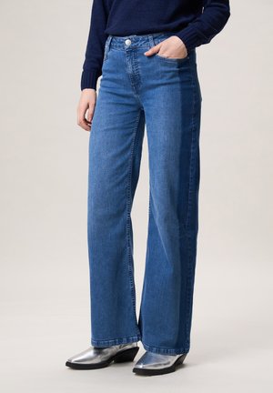 Persoon draagt een high-waist, tweekleurige blauwe wijde jeans met een voorzak en zilveren enkellaarsjes met puntige neus.