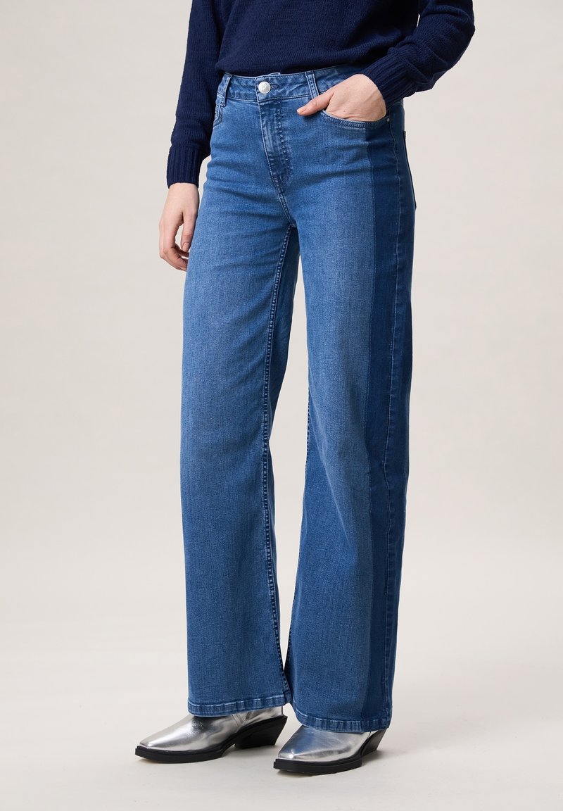 Persoon draagt een high-waist, tweekleurige blauwe wijde jeans met een voorzak en zilveren enkellaarsjes met puntige neus.