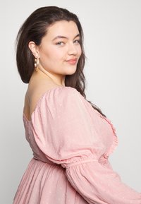 Vestido rosa claro com tecido texturizado, mangas bufantes e decote com folhos. Grandes brincos de argola dourada complementam o visual.