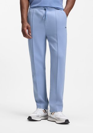 Uomo che indossa pantaloni da jogging azzurri chiaro con cuciture frontali e sneakers bianche, in piedi con le mani nelle tasche contro uno sfondo bianco.