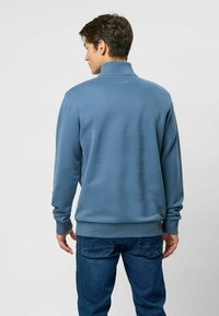 Kék pamut pulóver magas gallérral, bordázott mandzsettákkal és derékpánttal. Simaszerű textúrával és laza szabással rendelkezik, farmerek felett viselve.