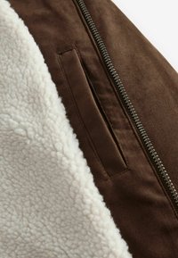 Giacca in suede marrone con una fodera in pile testurizzato di colore crema, dotata di una cerniera in metallo e di una tasca laterale all'esterno.