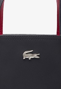 Sac à main noir texturé featuring un logo en crocodile argenté proéminent. L'intérieur est doublé de matériau rouge. Deux poignées robustes incluses.
