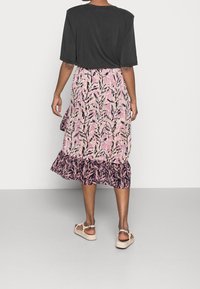 Mujer vestida con una blusa negra de manga corta, una falda midi con estampado en rosa y negro, y sandalias beige, de pie contra un fondo liso.