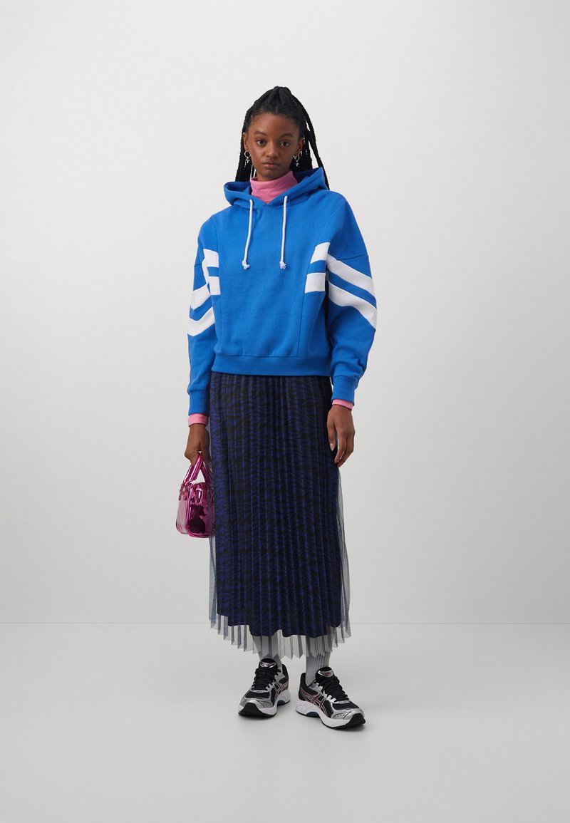 Blauwe hoodie met witte strepen, gedragen over een lange, gemusterde rok. Houdt een kleine roze handtas vast; draagt zwarte sneakers.