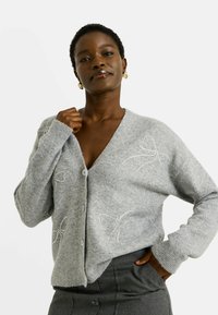 Cardigan en tricot gris avec un col en V, fermeture à boutons et motifs de nœuds en strass décoratifs sur les côtés. Texture douce et coupe décontractée.